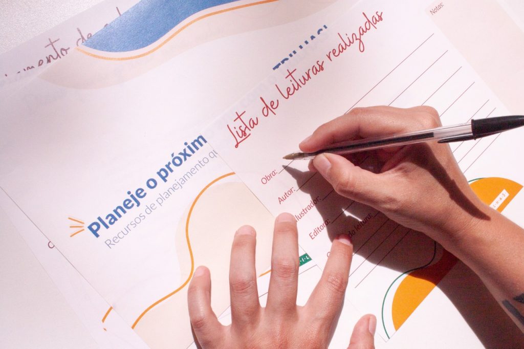 Duas mãos brancas escrevem com caneta sobre planejamento impresso